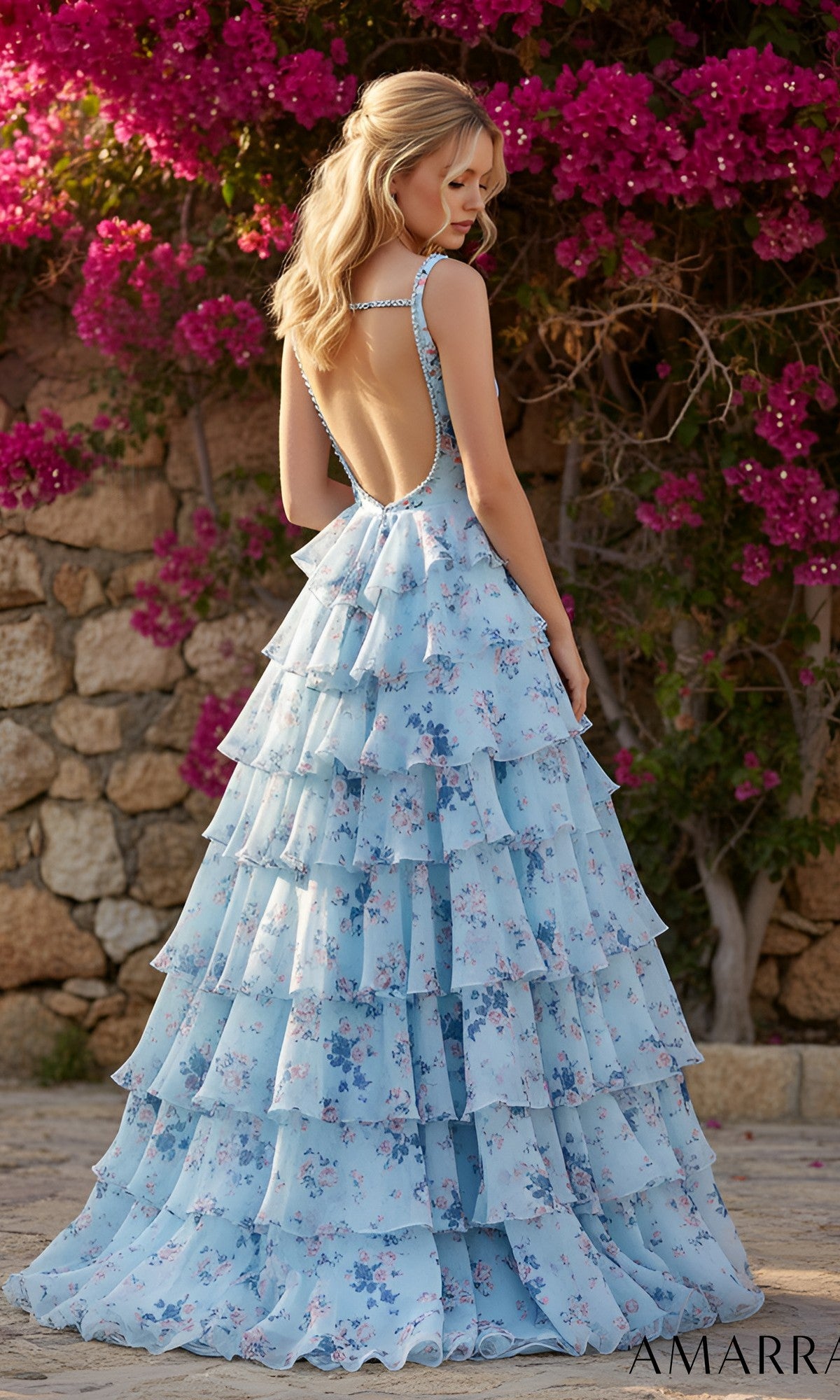 A woman stands outdoors in front of a stone wall and pink bougainvillea, wearing the Amarra 89613 Long Prom Dress—a backless, light blue floral-printed tiered gown with a flowing chiffon skirt.
